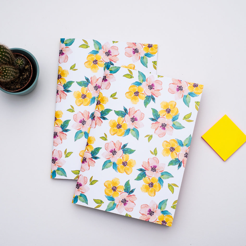Two floral notebooks on a light gray surface with a yellow post-it note and small plant.