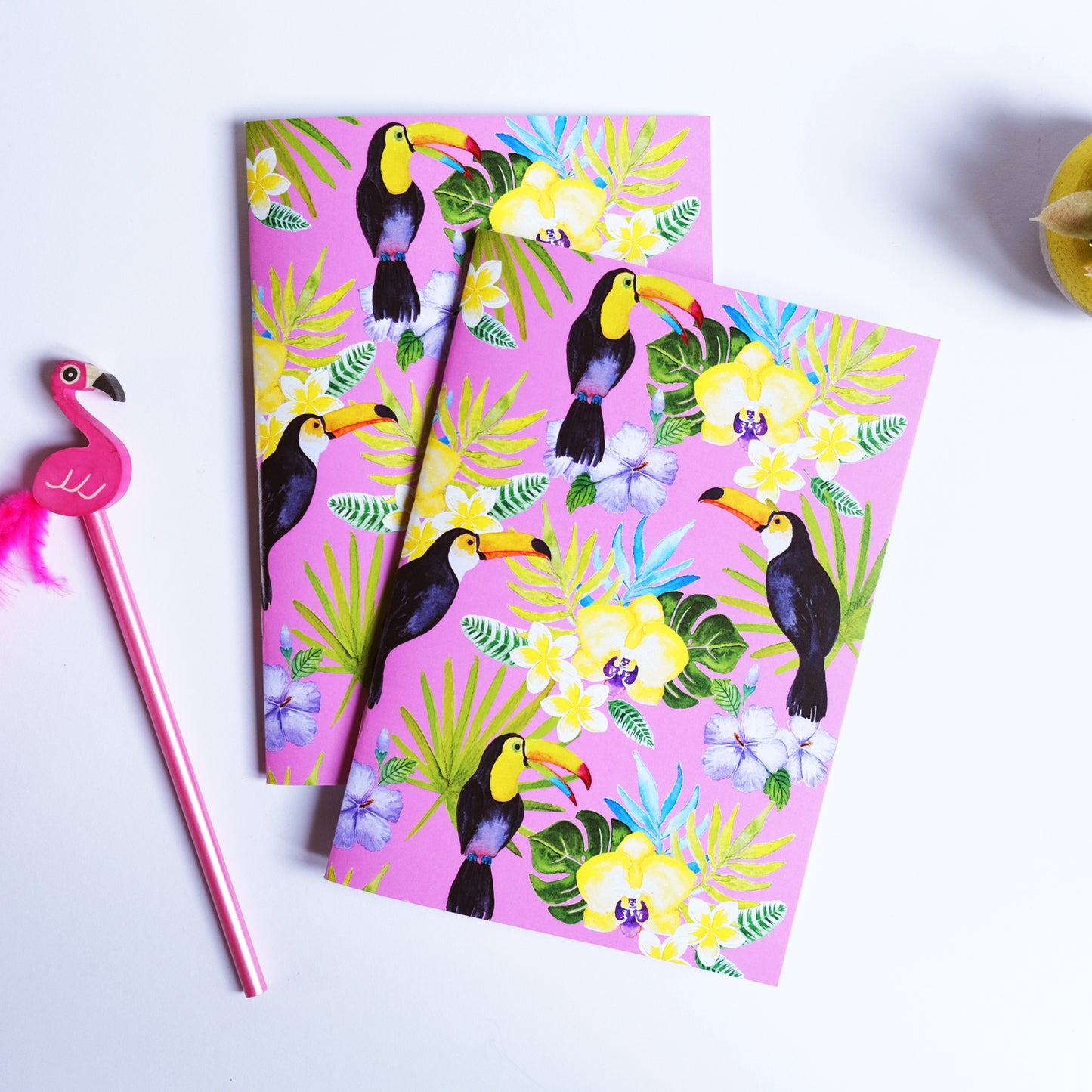 Two notebooks with a tropical bird design on a pink background, accompanied by a pink flamingo pen.