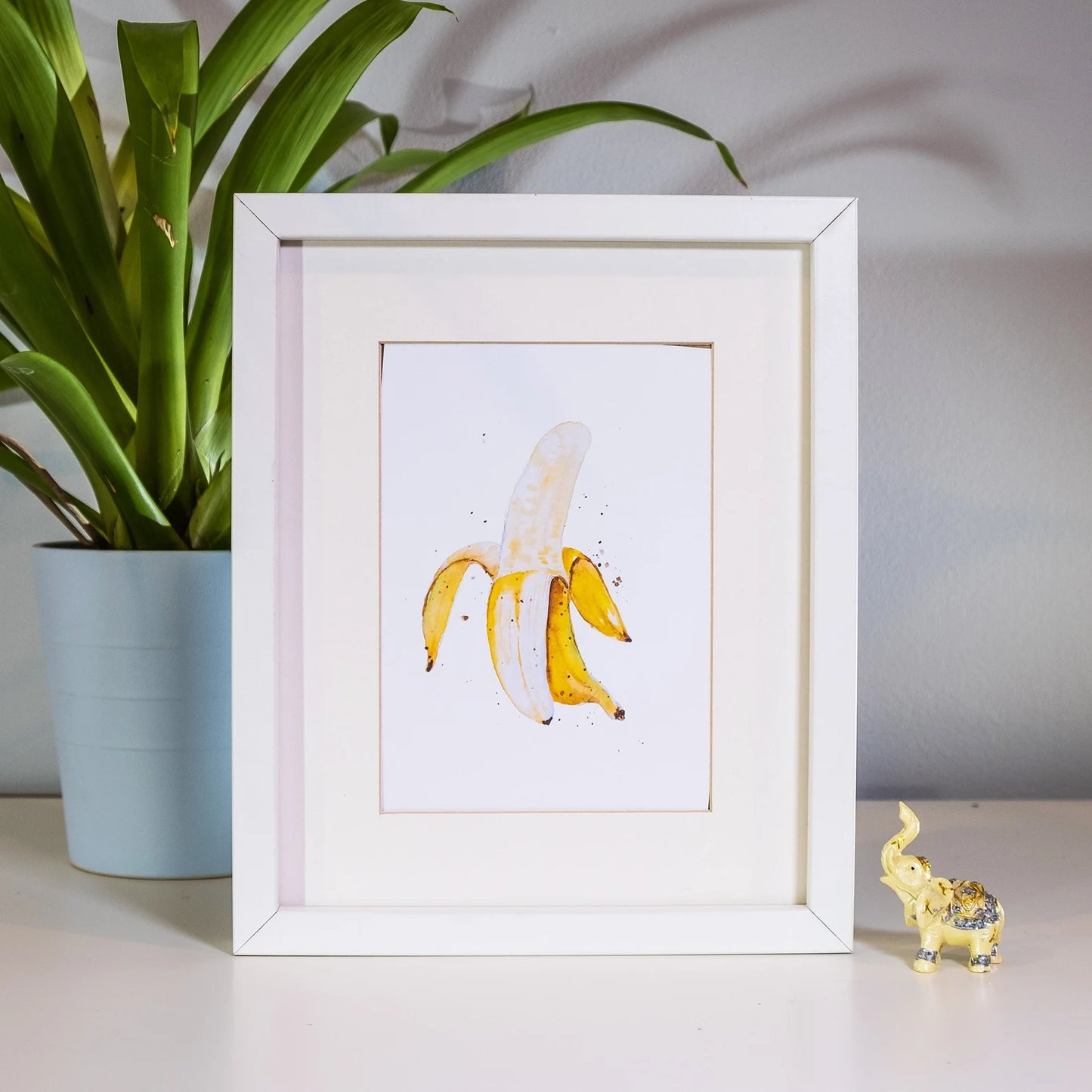 Framed artwork of a banana peel with a plant and small figurine in the background