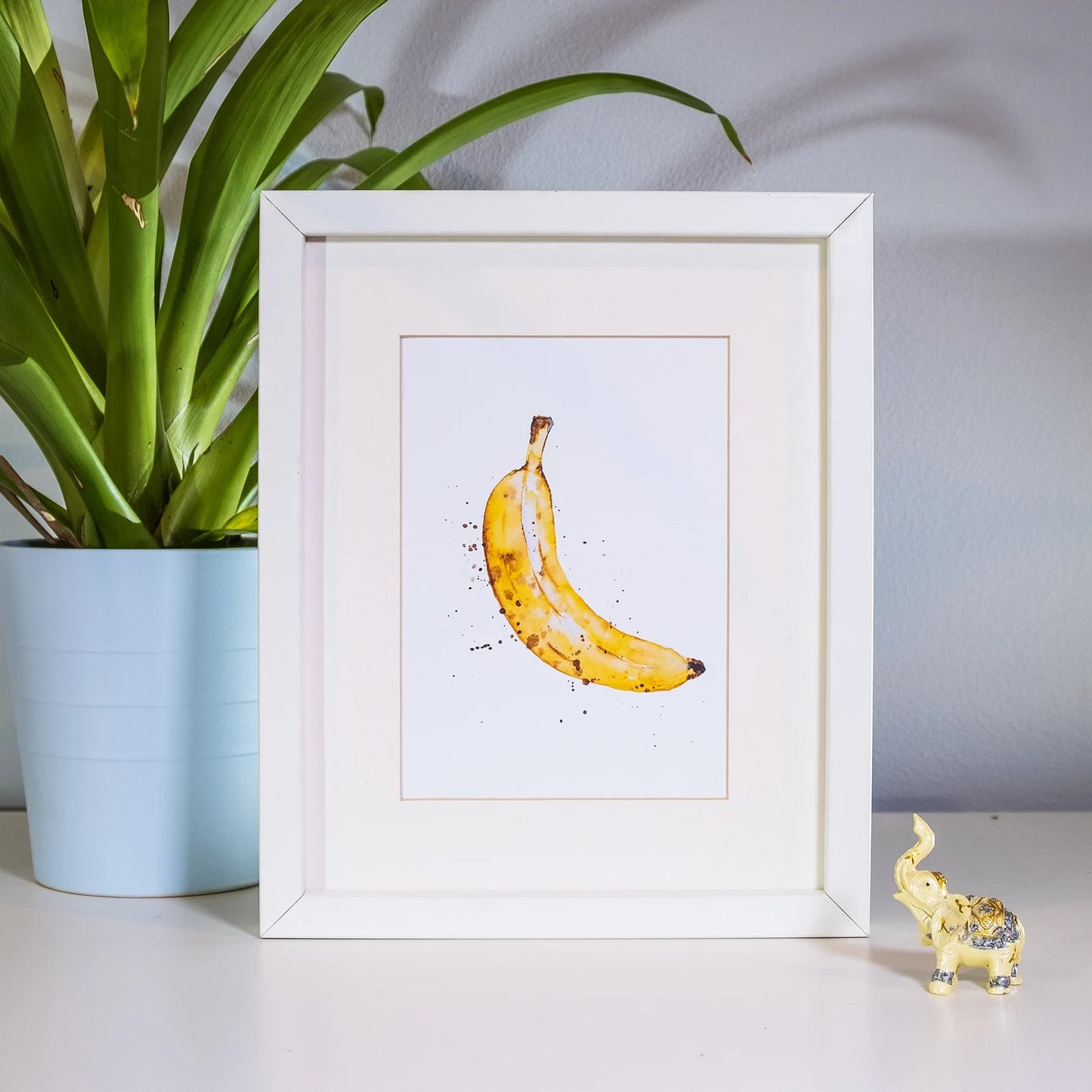 Framed artwork of a banana on a white surface with a plant and decorative object in the background.
