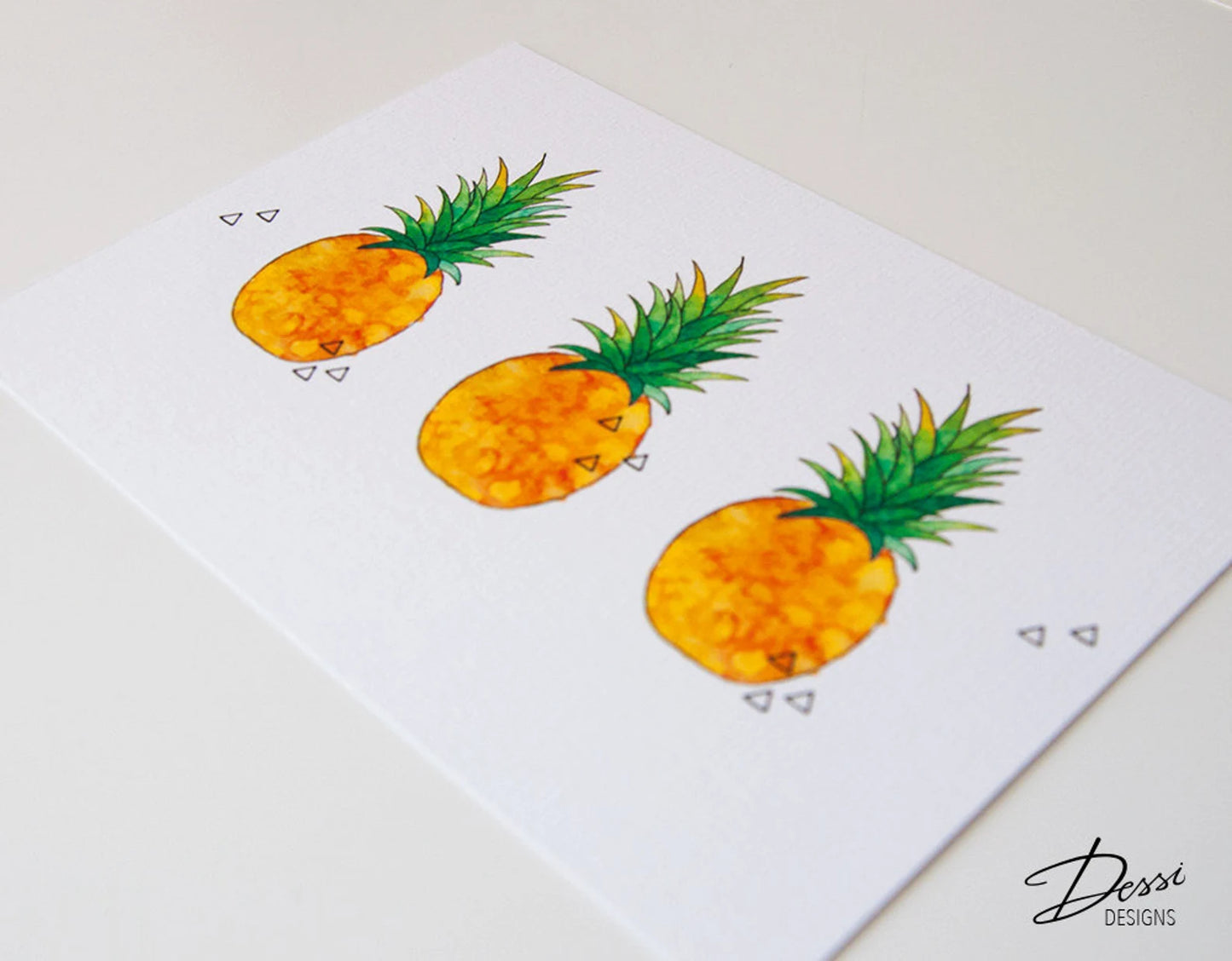 Three cartoon-style pineapples on a white card with 'Dessi Designs' branding.