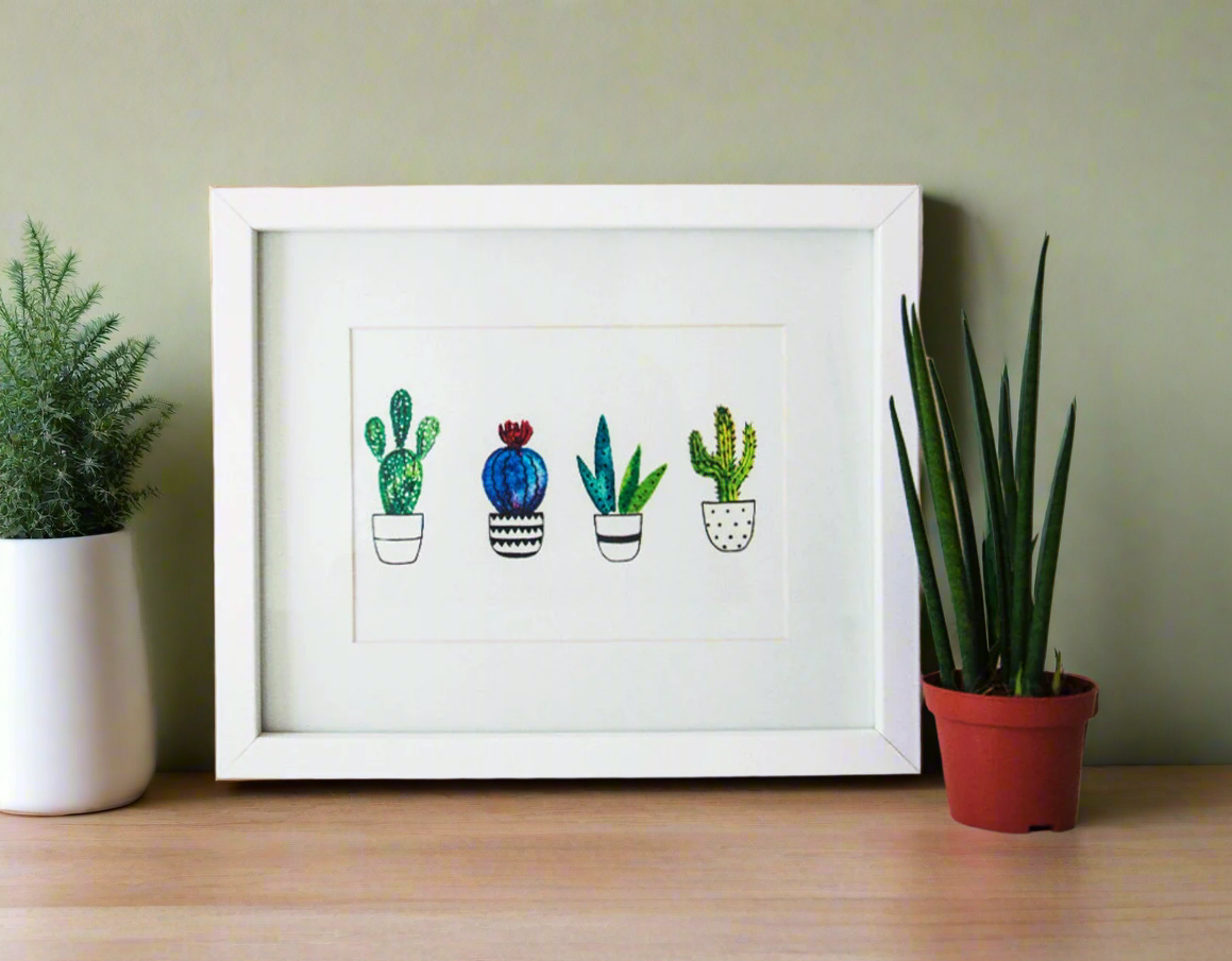 Framed artwork of cacti on a wooden surface with plants around