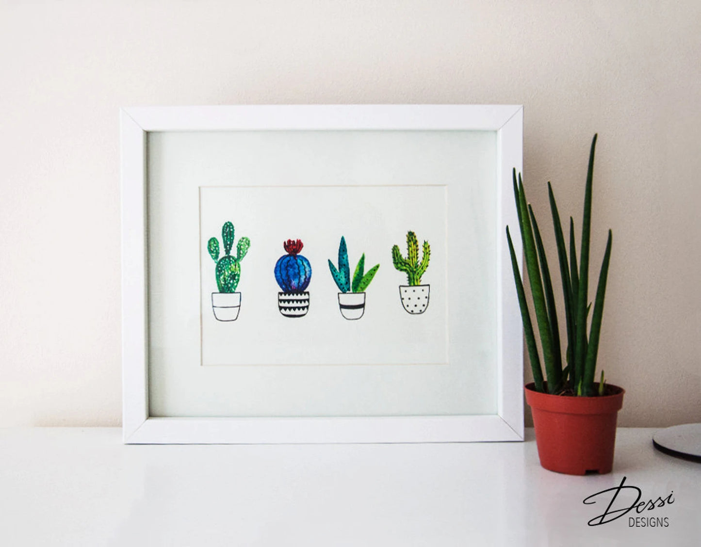 Framed artwork of cacti on a white surface with a plant in the background
