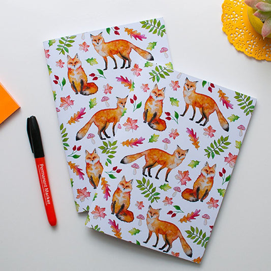 Two notebooks with fox and leaf pattern on a white surface with a pen and doily.