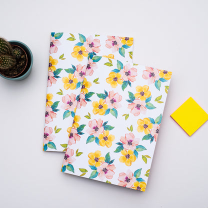 Two floral notebooks on a light gray surface with a yellow post-it note and small plant.