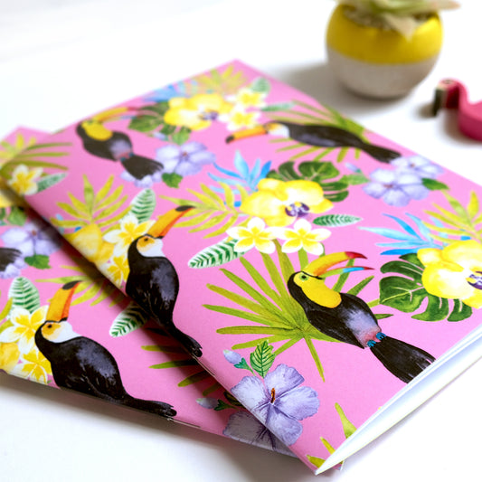 Two notebooks with toucan and floral design on a pink background