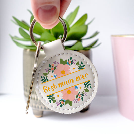 White faux leather Keychain with 'Best mum ever' text and floral design held by a hand.