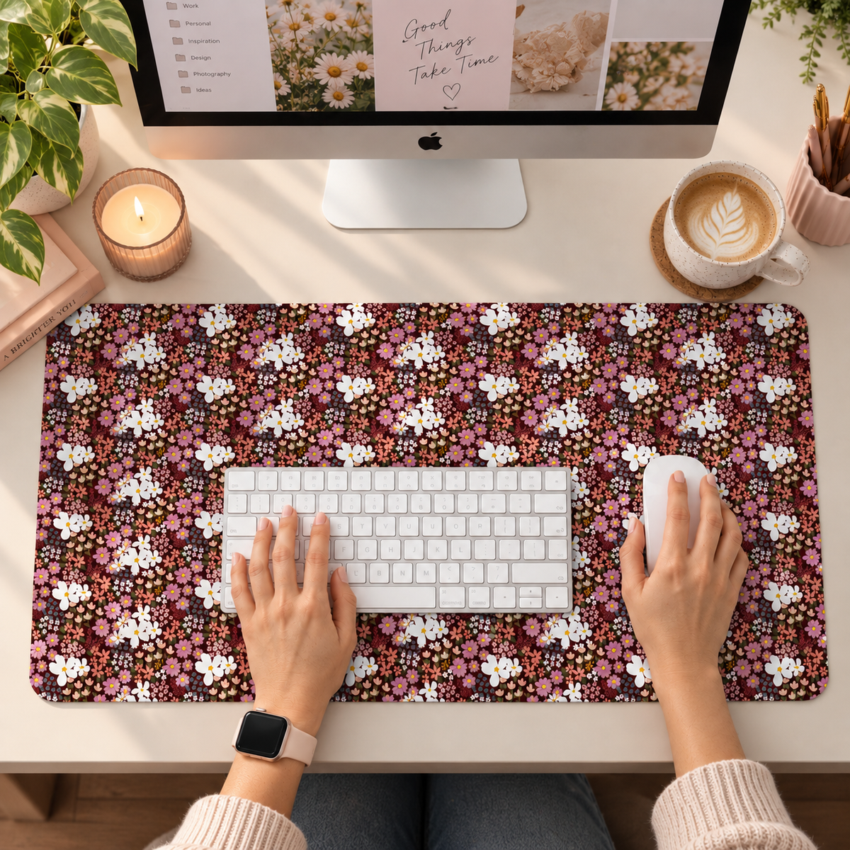 A top-down view of a desk setup showing a large rectangular mouse pad with small pink, white, and burgondy floral pattern. Hands are resting on a white keyboard and using a white mouse on the pad, with an iMac, candle, coffee cup, and plants visible in the scene.