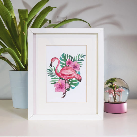 Framed artwork of a pink flamingo with flowers and leaves on a table with a plant and decorative glass ball in the background.
