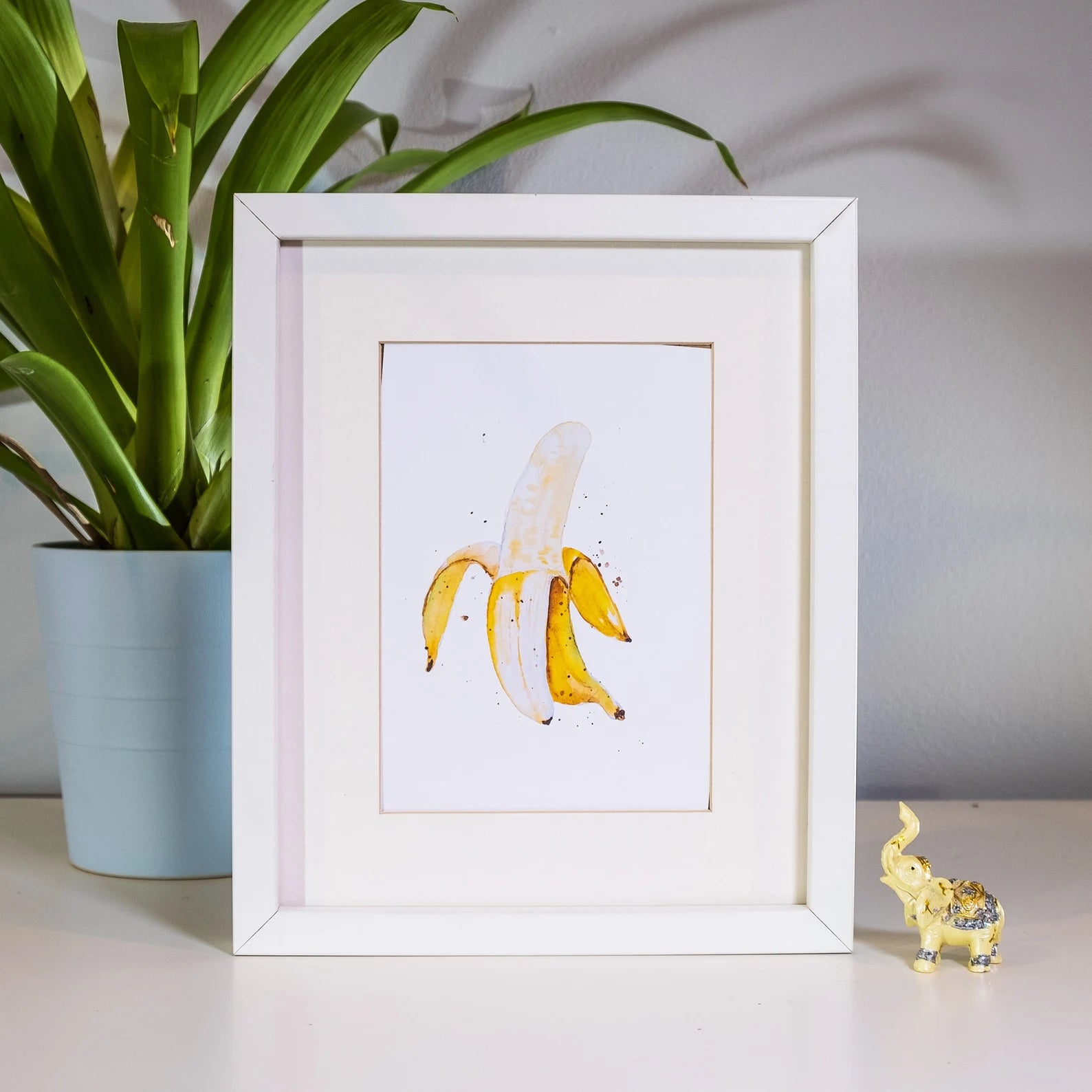 Framed artwork of a banana peel with a plant and small figurine in the background