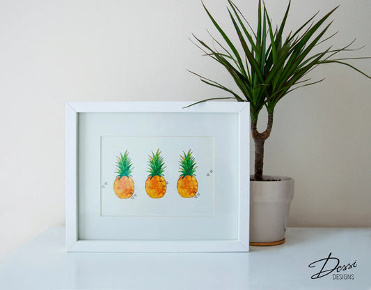 Framed artwork of three pineapples next to a potted plant on a white surface.