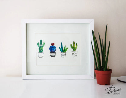 Framed artwork of cacti on a white surface with a plant in the background