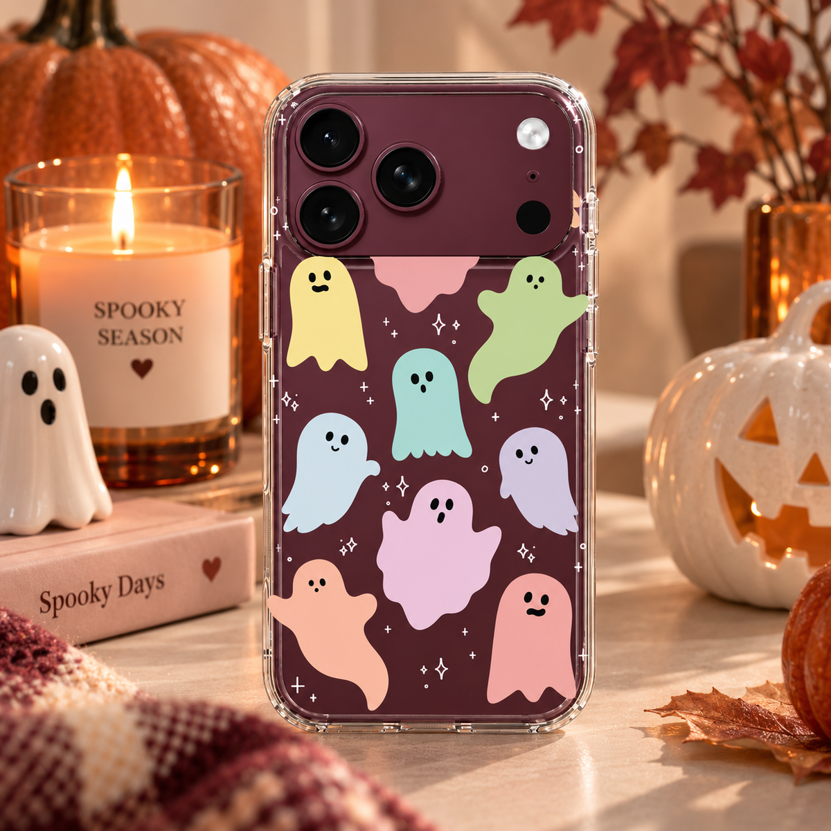 A clear phone case with a pattern of cute pastel ghosts, shown on a smartphone. The phone case is styled in a cozy, autumnal setting with a lit candle, a small white ghost figurine, a book, and a jack-o'-lantern pumpkin.